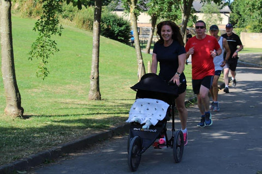 Woman running through a park, with a 3 wheeled running buggy with a baby inside, covered by a blanket. The woman is wearing running shorts and a tshirt, pink running shoes and behind her are several other people who are also running. 