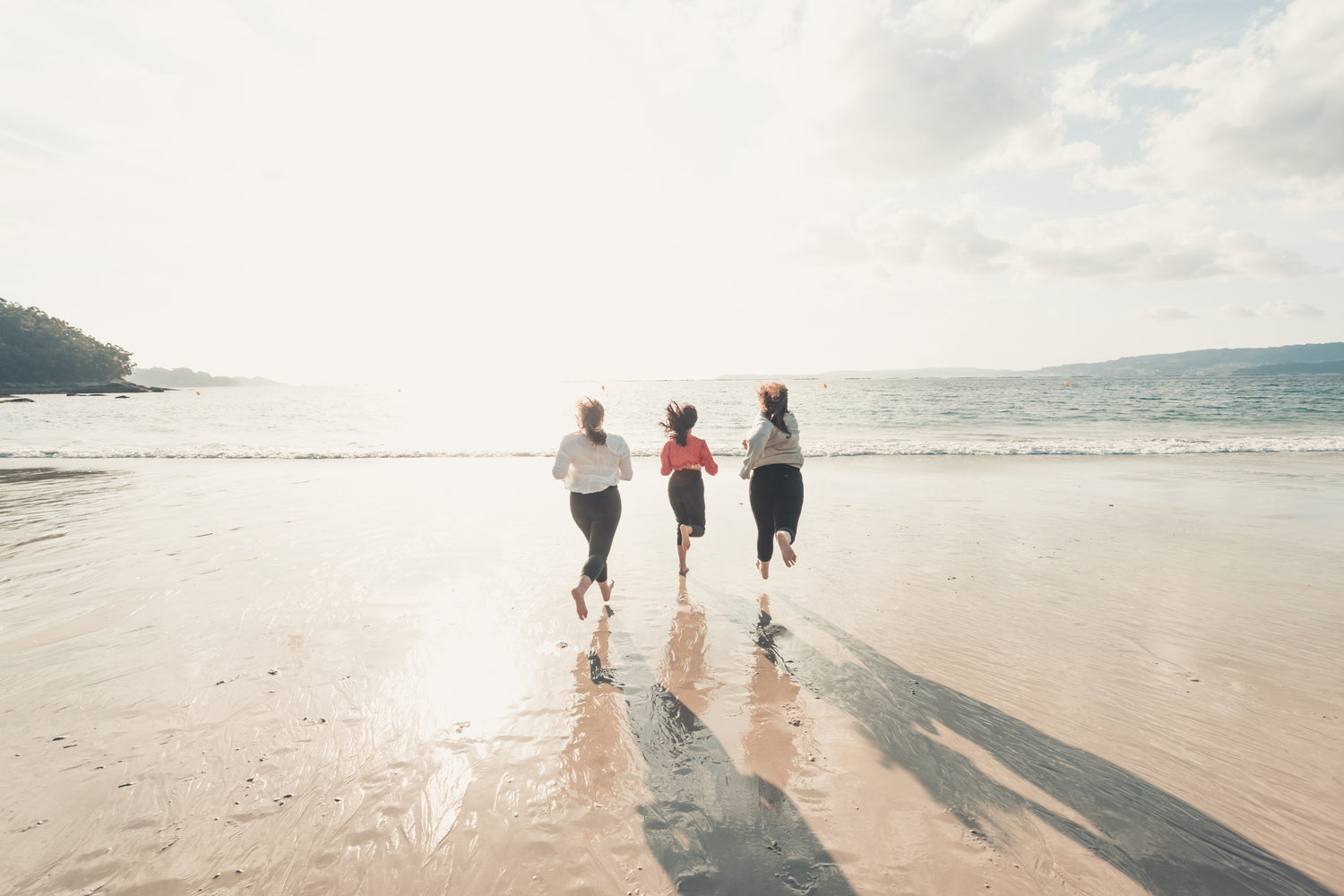 Three women running barefoot on a beach towards the ocean