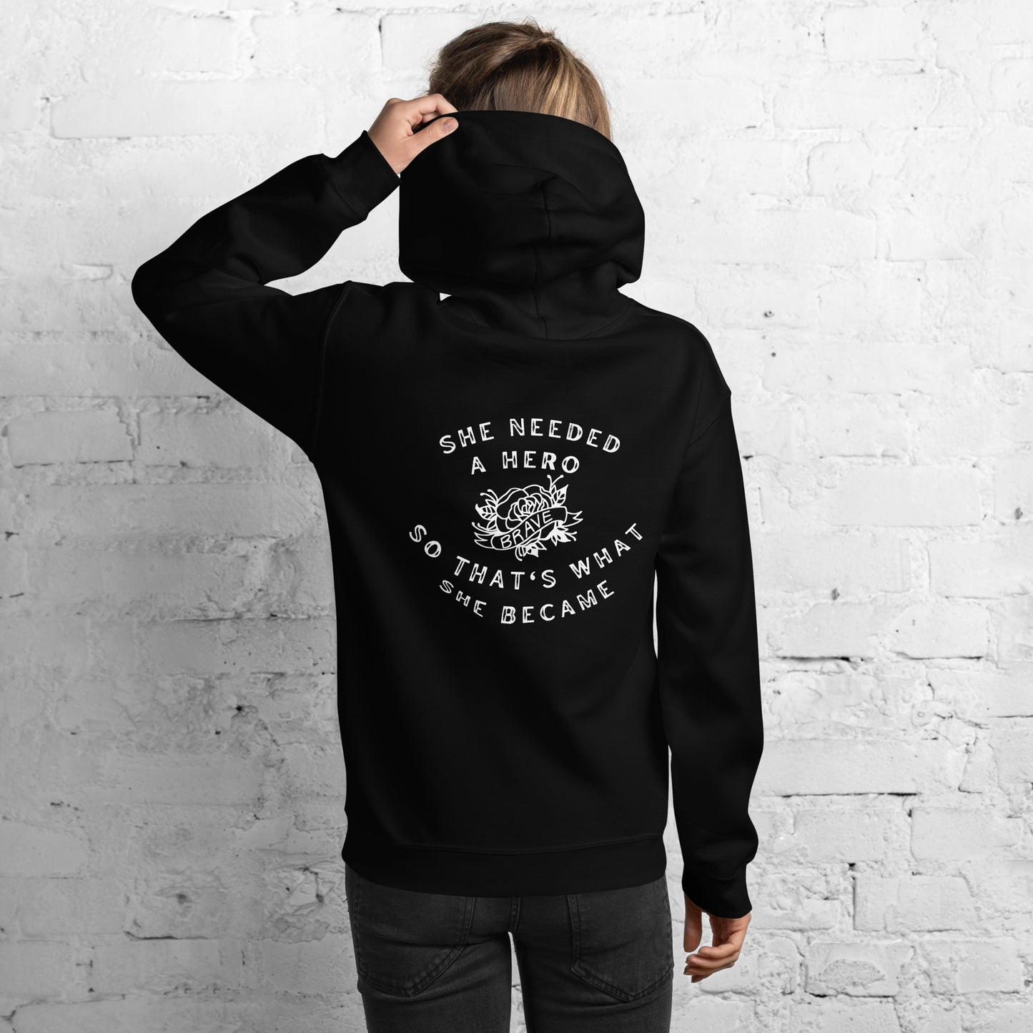 woman wearing a black hoodie shown from the back, with the words she needed a hero, so that's what she became wrapped around a tattoo style image of a rose with the word 'brave' in a scroll across it